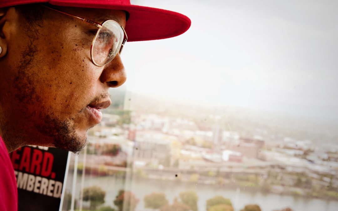 Close-up side profile of a man wearing a red hat and glasses looking out over a city skyline through a window, symbolizing focus and reflection.